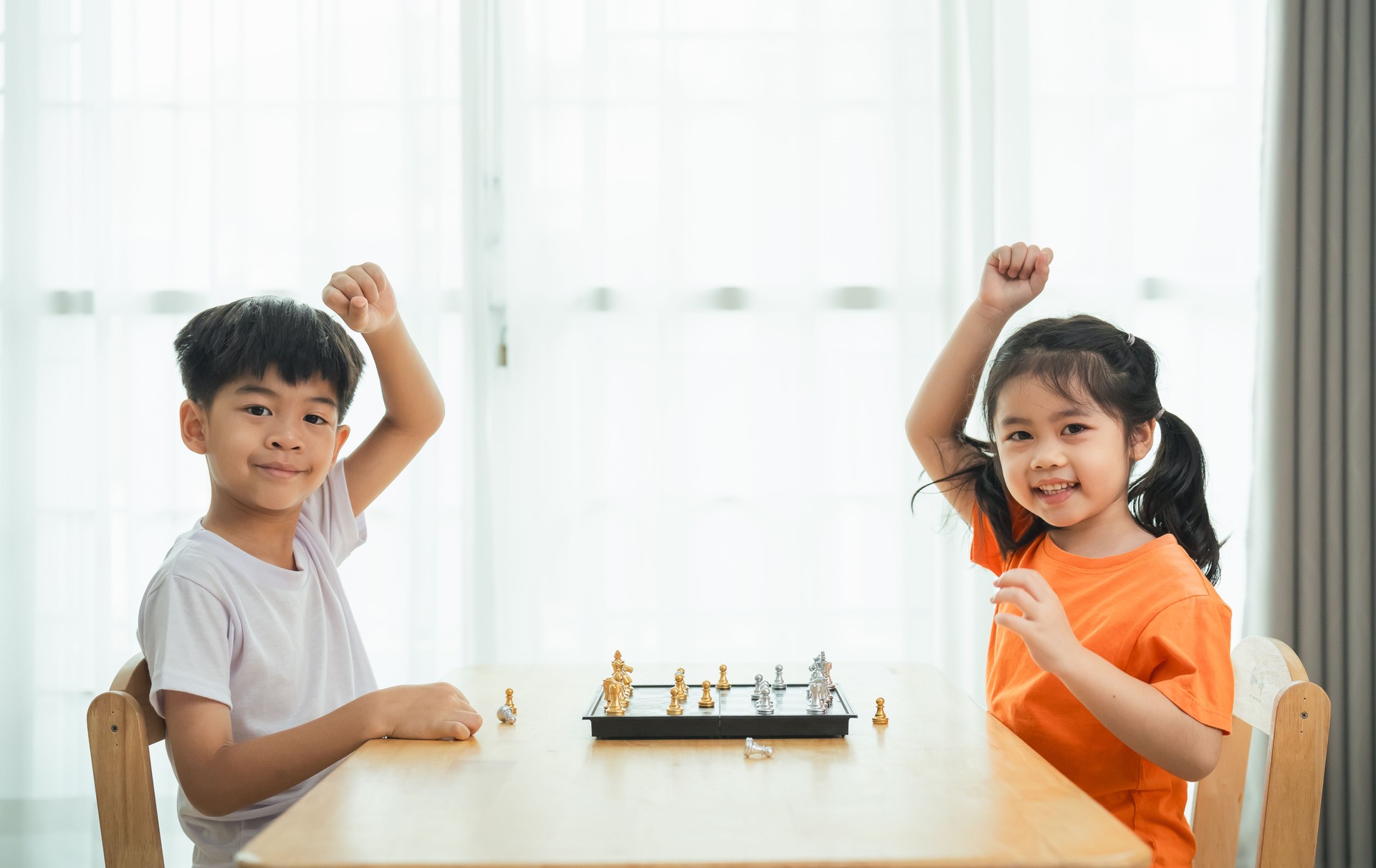 Two children playing a game of chess. One of the children is wearing an orange shirt. They are both smiling and seem to be enjoying the game